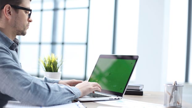 A male office worker is working on a laptop with a green screen.
