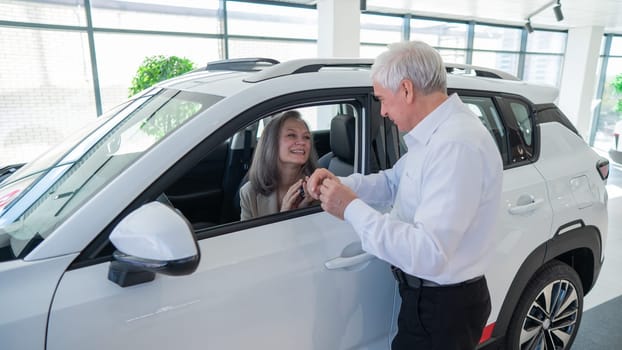 Mature Caucasian woman sits in a new car, an elderly man gives her the keys