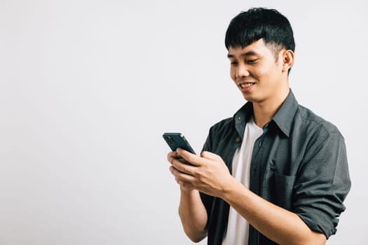 Smiling Asian teen enjoys online communication, typing a text message on his smart mobile phone. Studio portrait isolated on white, showcasing his enthusiasm for technology. Copy space available.