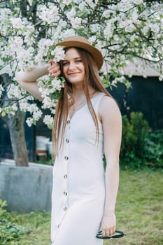 Beautiful young girl in white dress and hat in blooming Apple orchard. Blooming Apple trees with white flowers