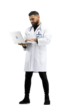 The doctor, in full height, on a white background, uses a laptop.