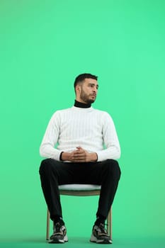 A man, full-length, on a green background, sitting on a chair.