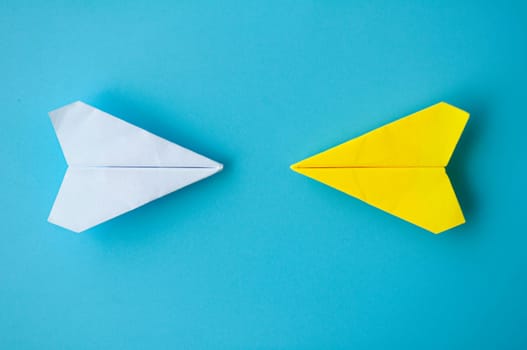 Top view of a white and yellow paper airplanes origami flying towards each other on blue background.