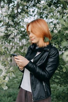 A red-haired teenage girl with headphones around her neck takes a selfie against the background of a flowering cherry tree. The time of spring flowering of cherry orchards