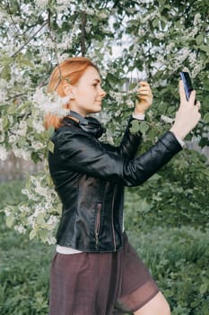 A red-haired teenage girl with headphones around her neck takes a selfie against the background of a flowering cherry tree. The time of spring flowering of cherry orchards.
