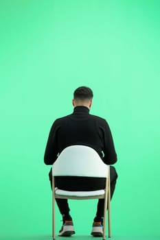 A man, full-length, on a green background, sitting on a chair.