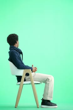 A man, full-length, on a green background, sitting on a chair.