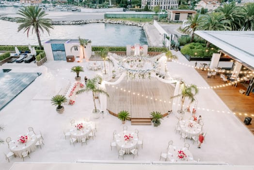 Round festive tables stand around a canopied stage decorated with glowing garlands by the sea. High quality photo