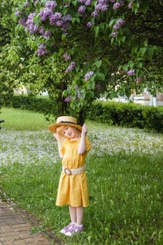 A little girl in a yellow dress and straw hat wearing a bouquet of lilacs. A walk in a spring park, blossoming lilacs