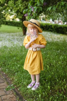 A little girl in a yellow dress and straw hat wearing a bouquet of lilacs. A walk in a spring park, blossoming lilacs