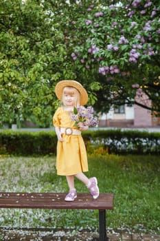 A little girl in a yellow dress and straw hat wearing a bouquet of lilacs. A walk in a spring park, blossoming lilacs