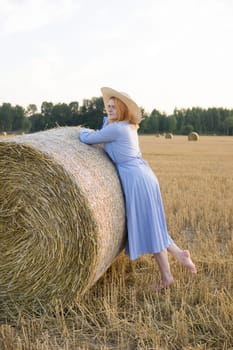 A red-haired woman in a hat and a blue dress walks in a field with haystacks