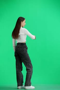 A woman, full-length, on a green background, looks at her watch.