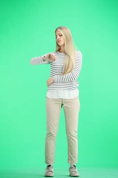A woman, full-length, on a green background, looks at her watch.