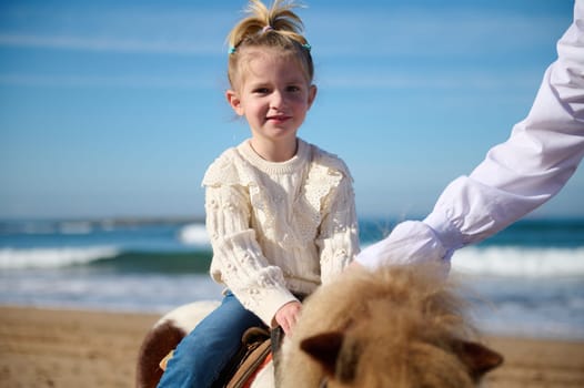 Happy Caucasian little child girl smiling looking at camera, riding a horse pony on the beach, smiling looking at camera. People. Active healthy lifestyle. Children and animals. leisure activity