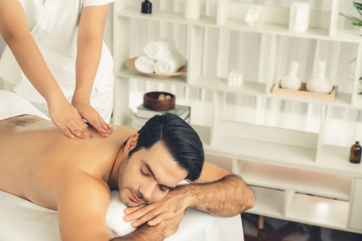 Caucasian man customer enjoying relaxing anti-stress spa massage and pampering with beauty skin recreation leisure in day light ambient salon spa at luxury resort or hotel. Quiescent