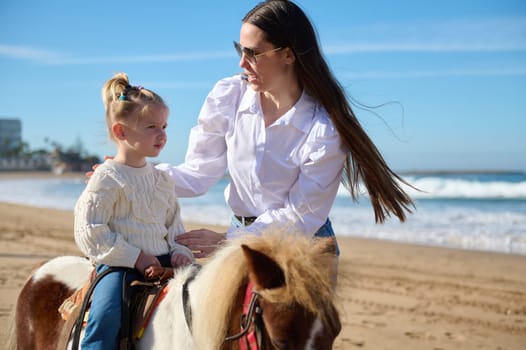 Beautiful Caucasian young woman, a loving caring mother teaching her lovely child to rise a horse pony. People. Nature. Animals. Happy carefree childhood. Leisure and hobbies.