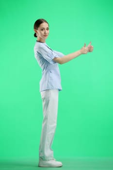 A female doctor, full-length, on a green background, shows a thumbs up.