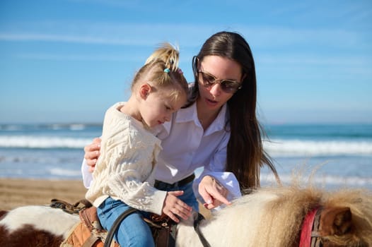Loving caring mother teaching her daughter to ride a horse pony on the beach. People. Active healthy lifestyle. Happy carefree childhood. Hobbies and leisure