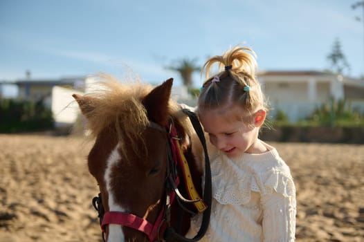 Adorable blonde little child girl stroking a pony, smiling and enjoying happy weekend outdoor. Children and love for animals