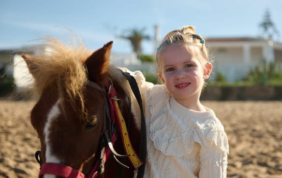 Caucasian cute kid girl hugging a little pony, smiling looking at camera, enjoying a happy family weekend outdoor. People, children, animals and nature concept. Weekend activities. Leisure and hobbies