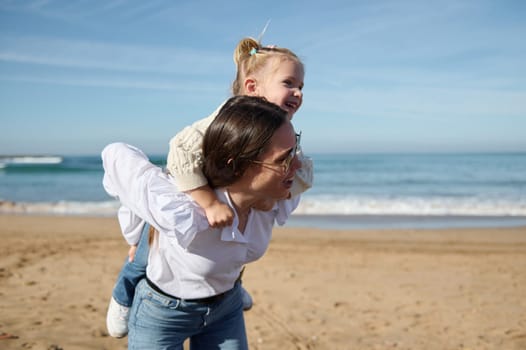 Happy young mother giving piggyback ride to her little daughter, enjoying walk together on the sandy beach. Family relationships. People. Active healthy lifestyle