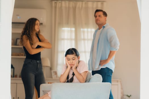 Annoyed and unhappy young girl sitting on sofa trapped in middle of tension by her parent argument in living room. Unhealthy domestic lifestyle and traumatic childhood develop to depression.Synchronos