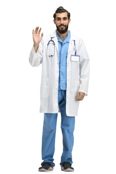 A male doctor, full-length, on a white background, waving his hand.