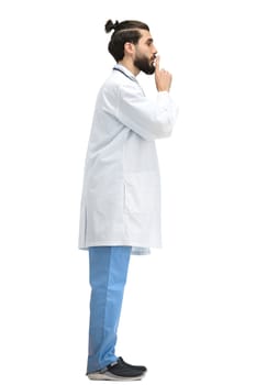 A male doctor, full-length, on a white background, shows a sign of silence.