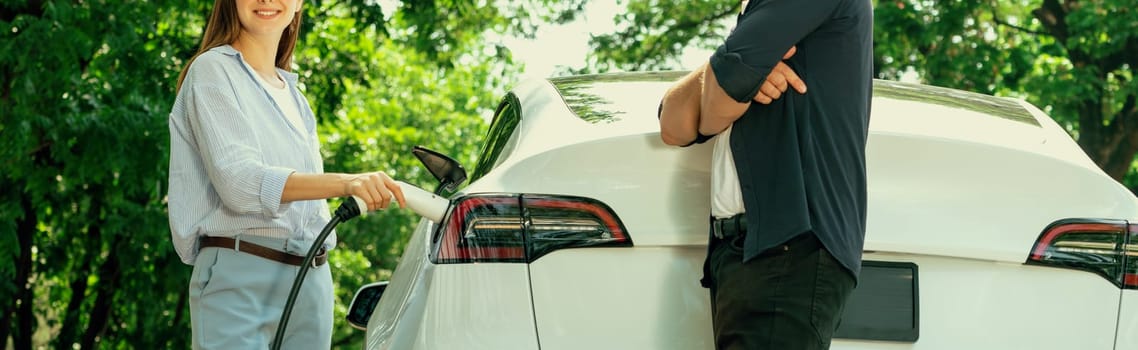 Focused EV car recharging electricity for battery on blurred background of lovey couple ruing their road trip travel by eco friendly electric car in national park and greenery forest on holiday. Exalt