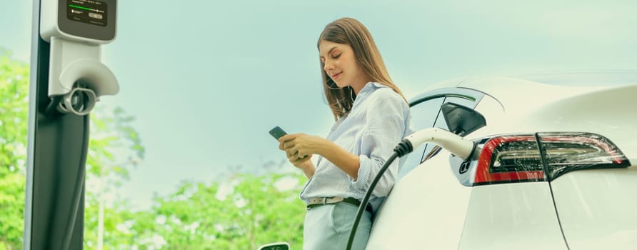 Young woman using smartphone online banking application to pay for electric car battery charging from EV charging station during vacation road trip at national park or summer forest. Panorama Exalt