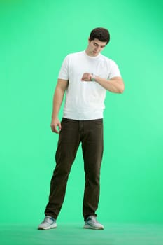 A man, full-length, on a green background, looks at his watch.