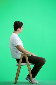 A man, full-length, on a green background, sitting on a chair.