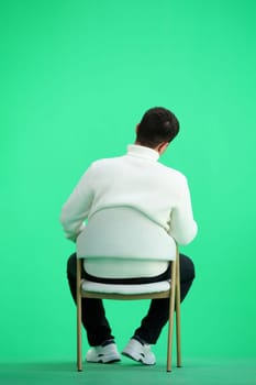 A man, full-length, on a green background, sitting on a chair.