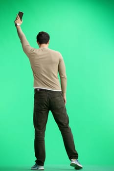 A man, full-length, on a green background, waving his phone.