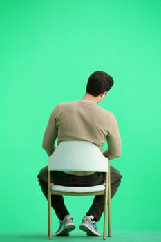 A man, full-length, on a green background, sitting on a chair.