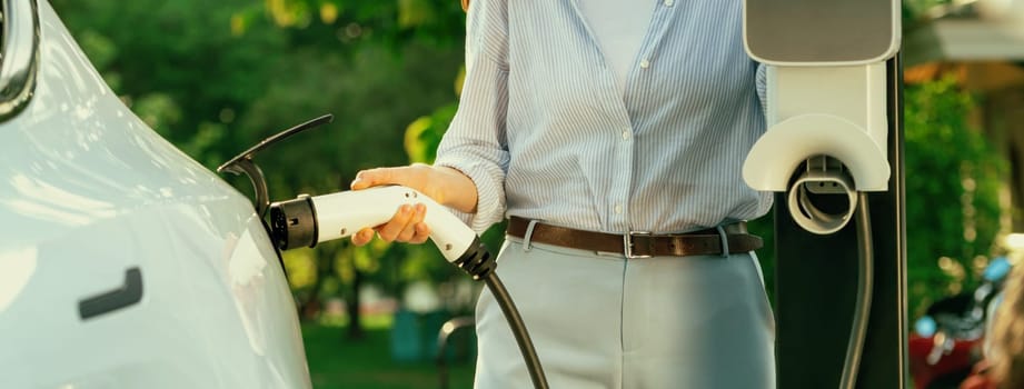 Young woman recharging battery for electric car during road trip travel EV car in natural forest or national park with sunrise lighting. Eco friendly travel during vacation and holiday. Panorama Exalt