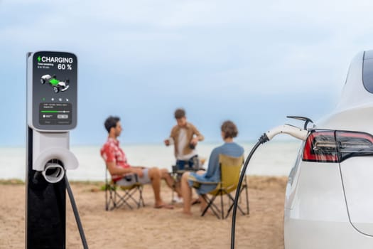 Alternative family vacation trip traveling by the beach with electric car recharging battery from EV charging station with blurred family enjoying the seascape background. Perpetual