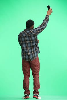 A man, full-length, on a green background, waving his phone.