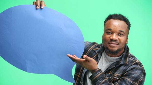 A man, close-up, on a green background, shows a blue comment sign.