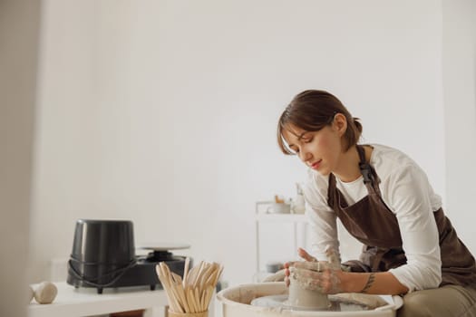 Concentrated female artisan in apron sitting on bench with pottery wheel and making clay pot