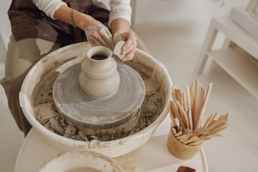 Female potter in apron making shape of clay vase on spinning pottery tool in ceramic workshop