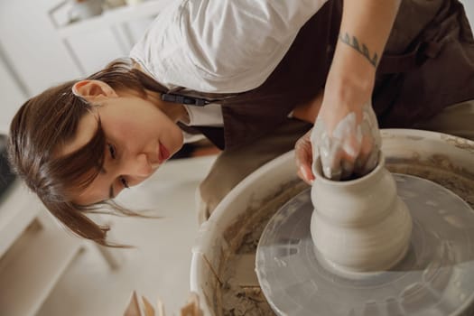 Concentrated female artisan in apron sitting on bench with pottery wheel and making clay pot