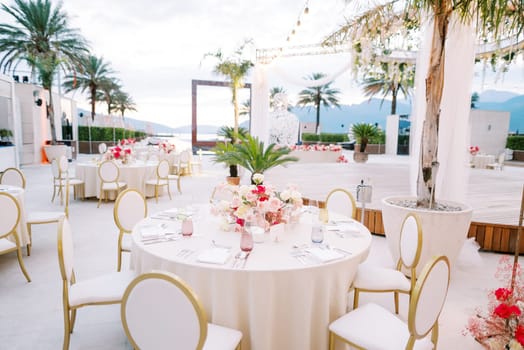 Festive tables with bouquets of flowers and white chairs stand near a stage decorated with garlands of light bulbs. High quality photo