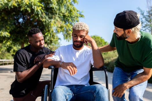 Multiracial friends consoling and comforting sad black male friend on a wheelchair in park outdoors. Friendship concept.