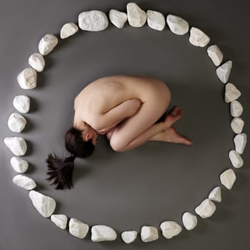 Nude woman lying in circle made of stones, on gray background