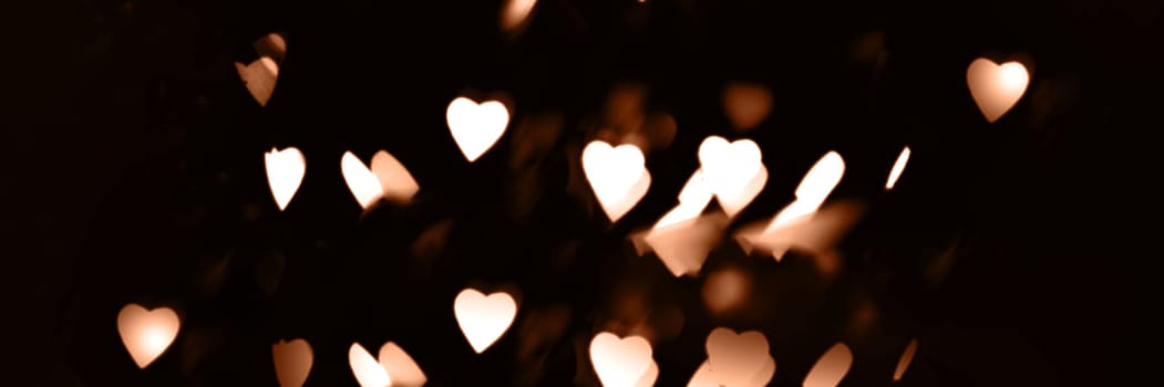 Blurry lights on a dark background. Festive bokeh background in the form of hearts for Valentine's Day. Soft focus. Image toned in color of the year 2024 - Peach Fuzz.