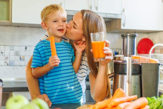 A joyful mom kisses her son, holding a carrot and fresh juice