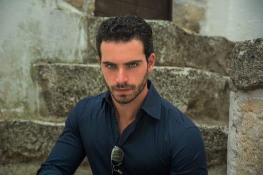 A man wearing a blue shirt is shown sitting down in the photograph. He appears relaxed and comfortable in his seated position. The background is simple and non-distracting, allowing the focus to be on the man in the blue shirt.