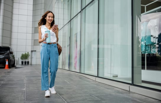 Female pedestrian with a tumbler mug and smartphone, engrossed in digital communication while walking on a city street